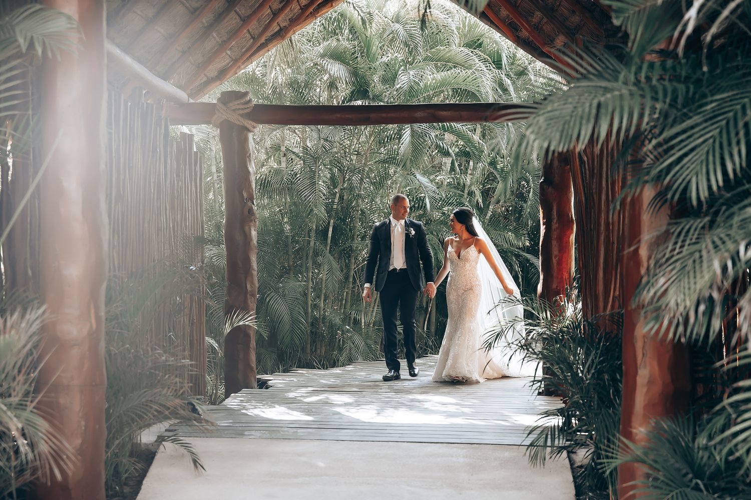 A couple walks through a tropical palm-lined pathway in wedding attire.