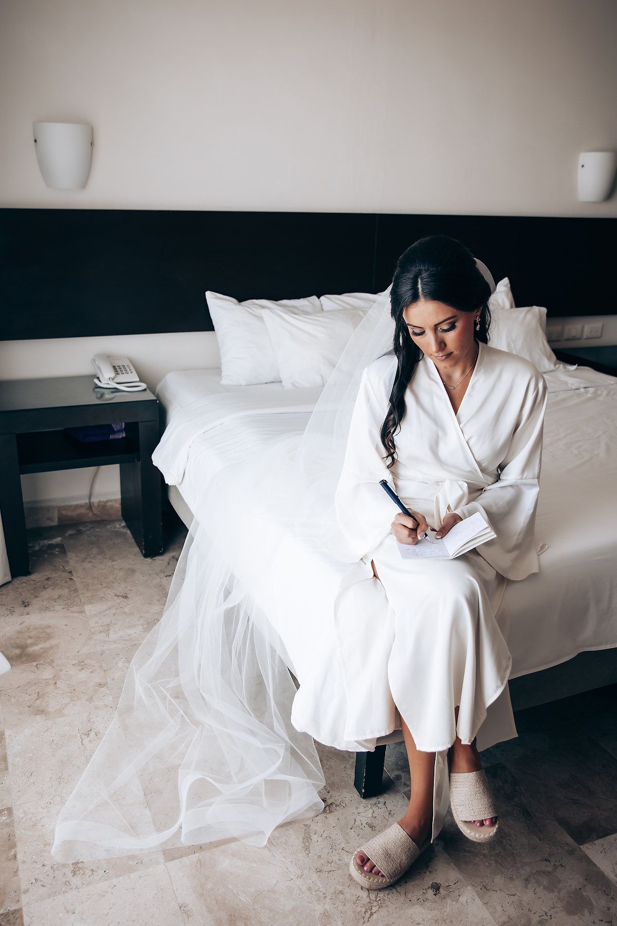 Person in white robe writing while sitting on bed with wedding dress hanging nearby.