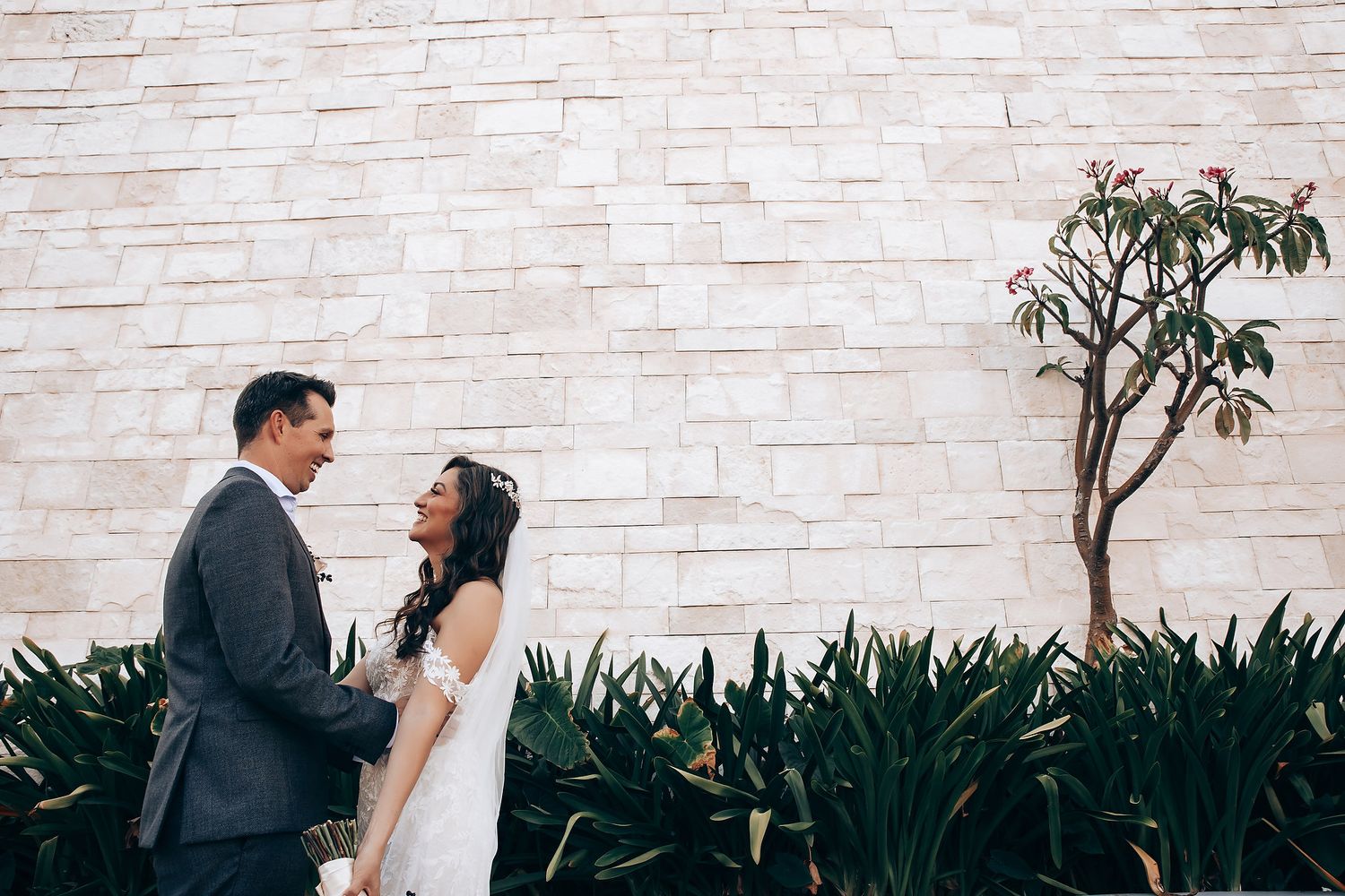 A romantic wedding couple poses against a white brick wall surrounded by lush tropical greenery.