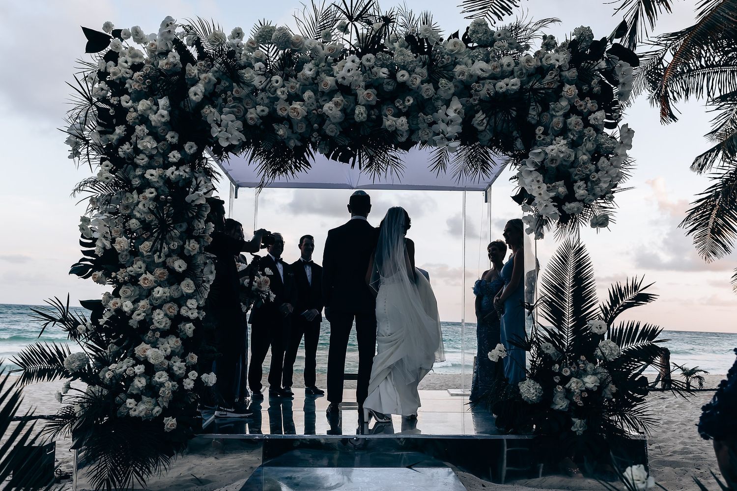 Silhouettes of wedding party gathered at sunset with tropical foliage and ocean view.