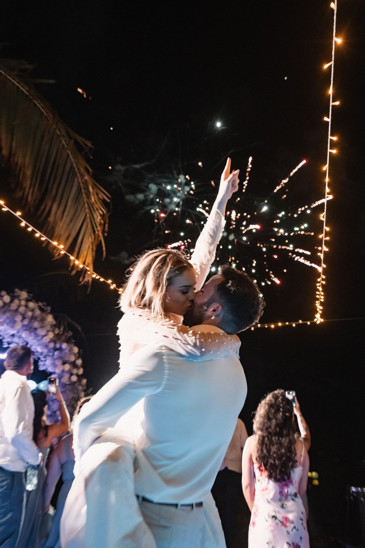 Groom lifting bride for romantic kiss with fireworks and string lights at outdoor night reception.