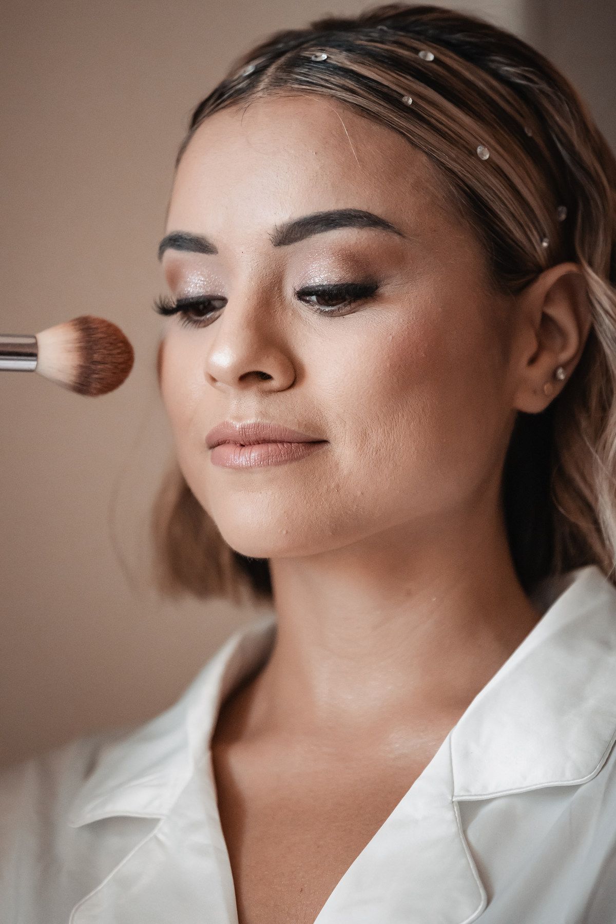 A makeup artist applying powder blush to a young woman with elegant eye makeup and hair accessories.