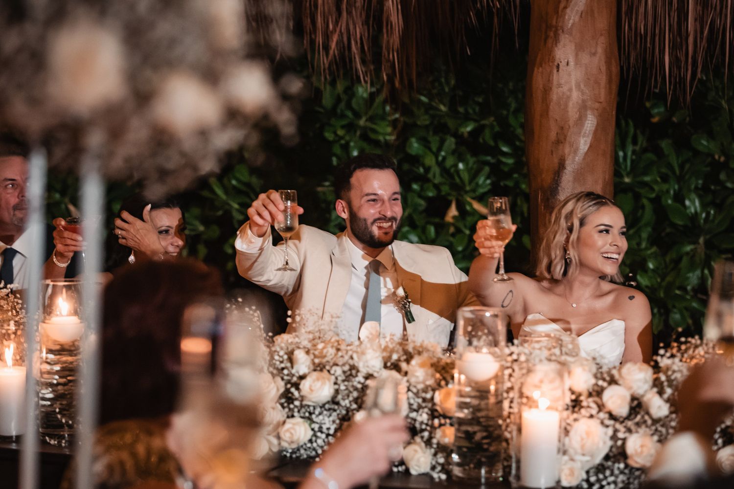 Newlyweds raise champagne glasses at candlelit sweetheart table adorned with white floral arrangements.