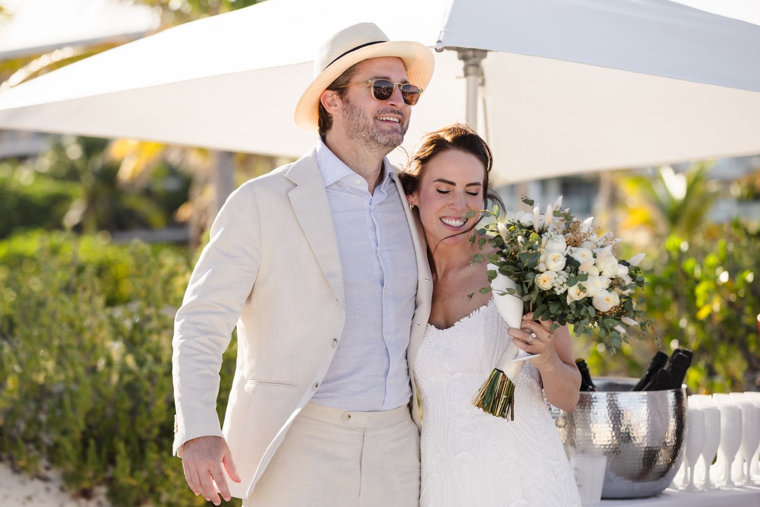 Happy couple in wedding attire smiling outdoors, bride holding white bouquet, groom in tan suit and hat.
