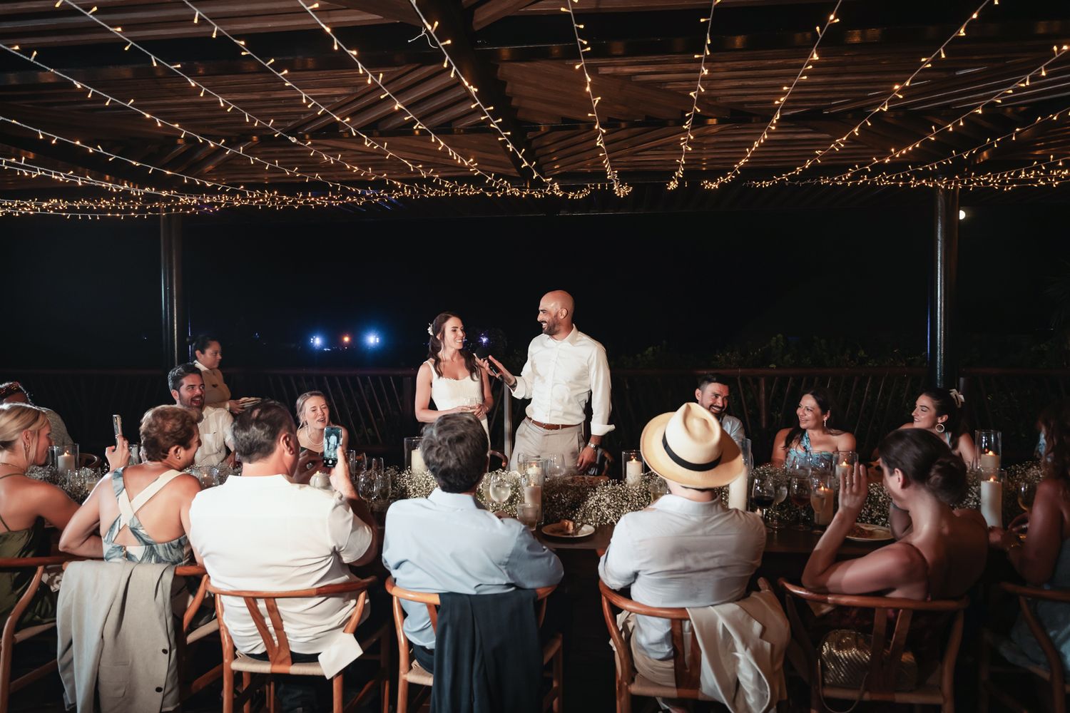 Couple giving a wedding speech at an outdoor reception under string lights with guests seated at tables.