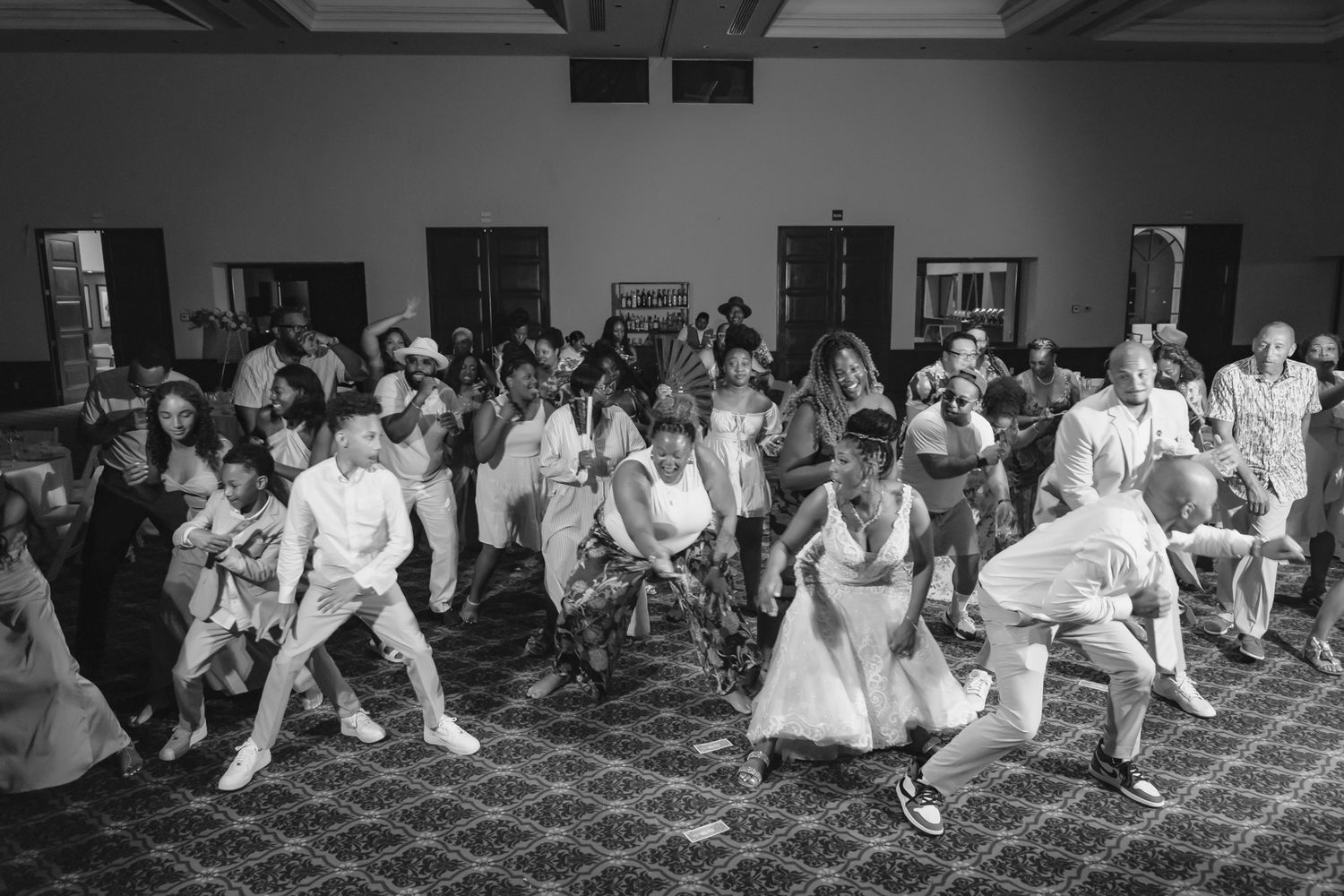 Black and white photo of wedding guests dancing joyfully at a reception in an elegant ballroom.
