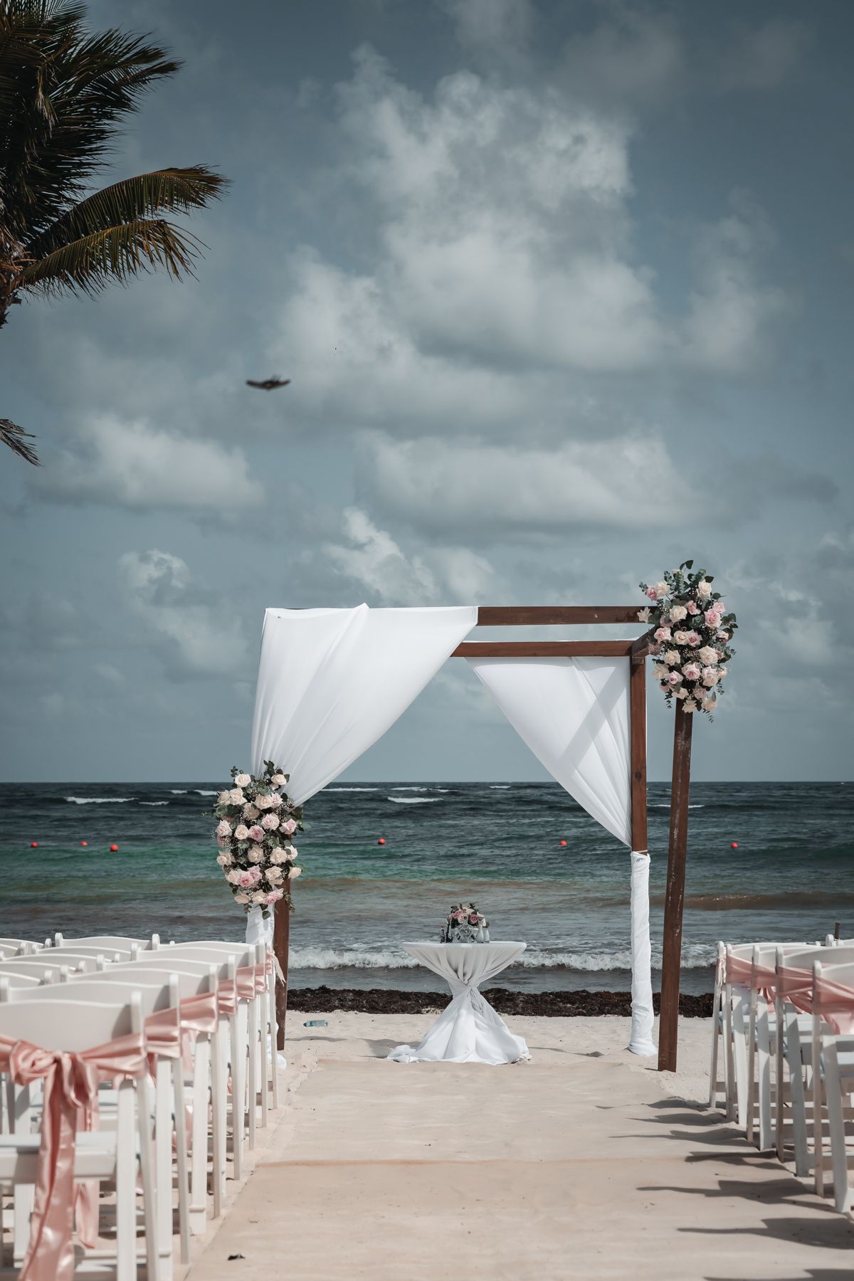 Elegant beachside wedding setup with white draped arch, pink floral arrangements, and white chairs along sandy aisle overlooking ocean.