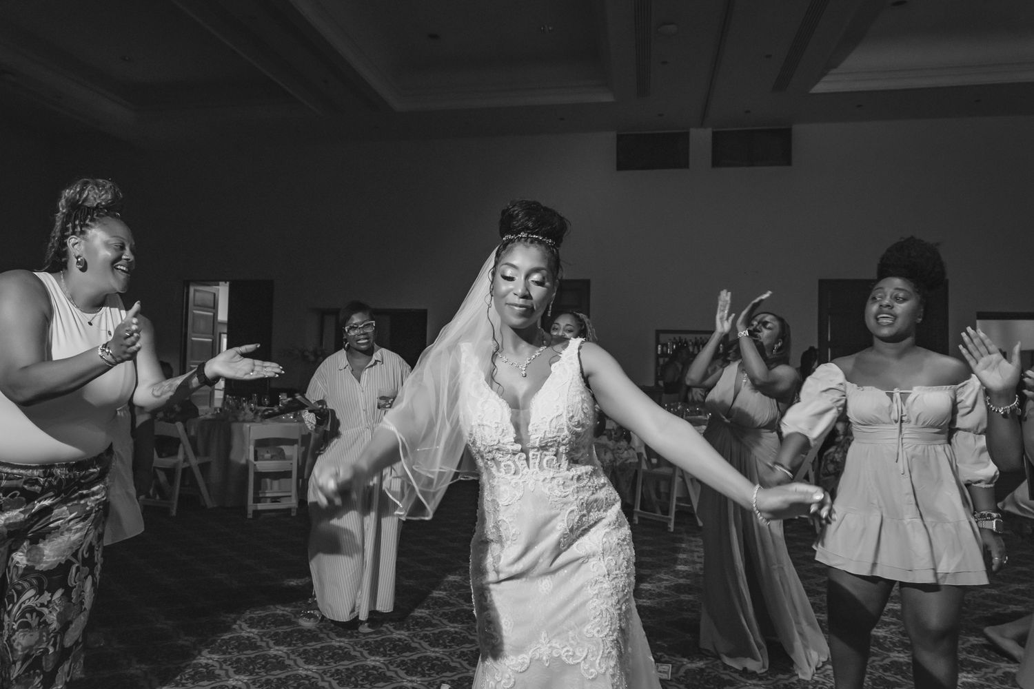 Bride in lace wedding gown and veil dancing joyfully with bridesmaids at wedding reception, black and white photo.