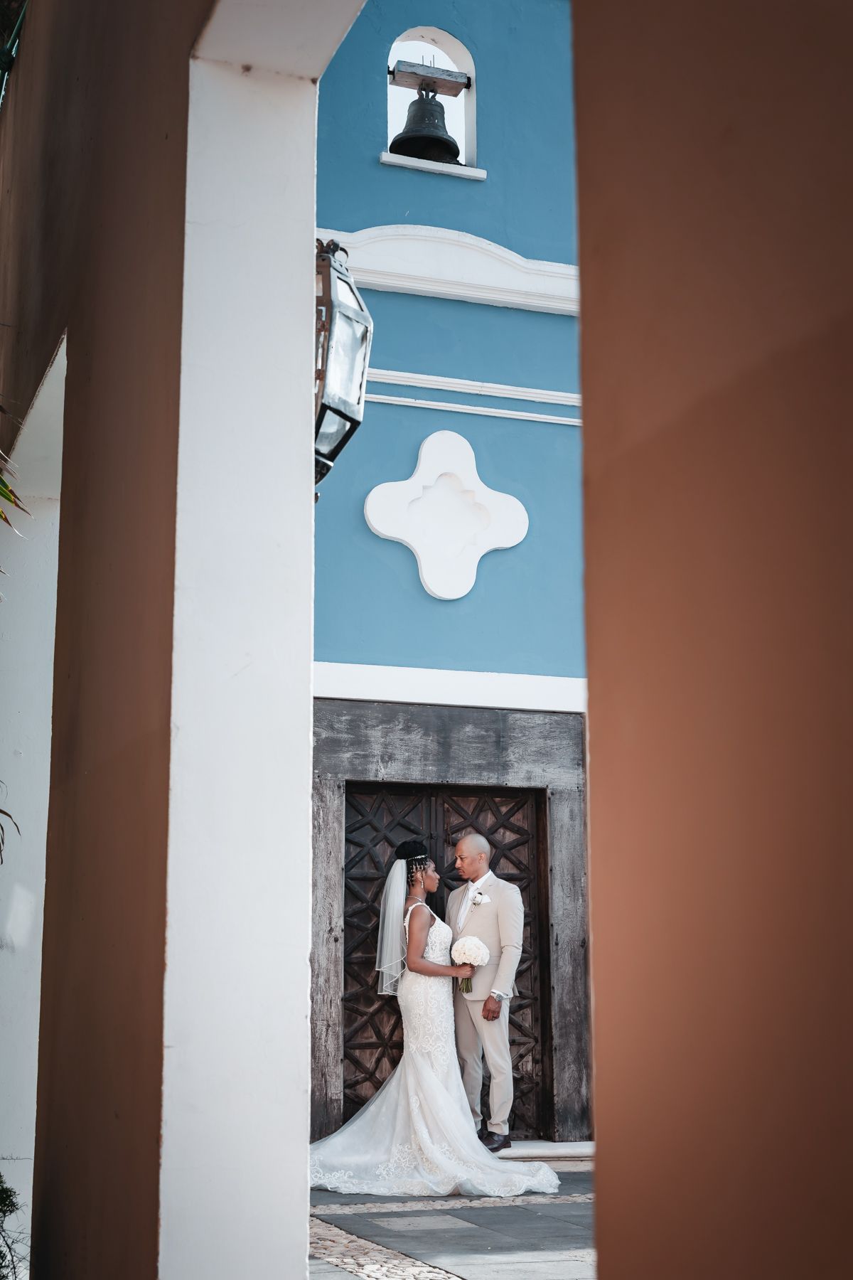 Bride and groom share a romantic moment in front of a blue church with a bell tower and ornate wooden doors.