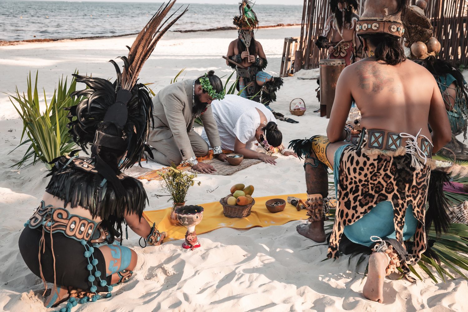 Indigenous ceremony on a white sand beach with participants in traditional Mayan attire and feathered headdresses performing a ritual.