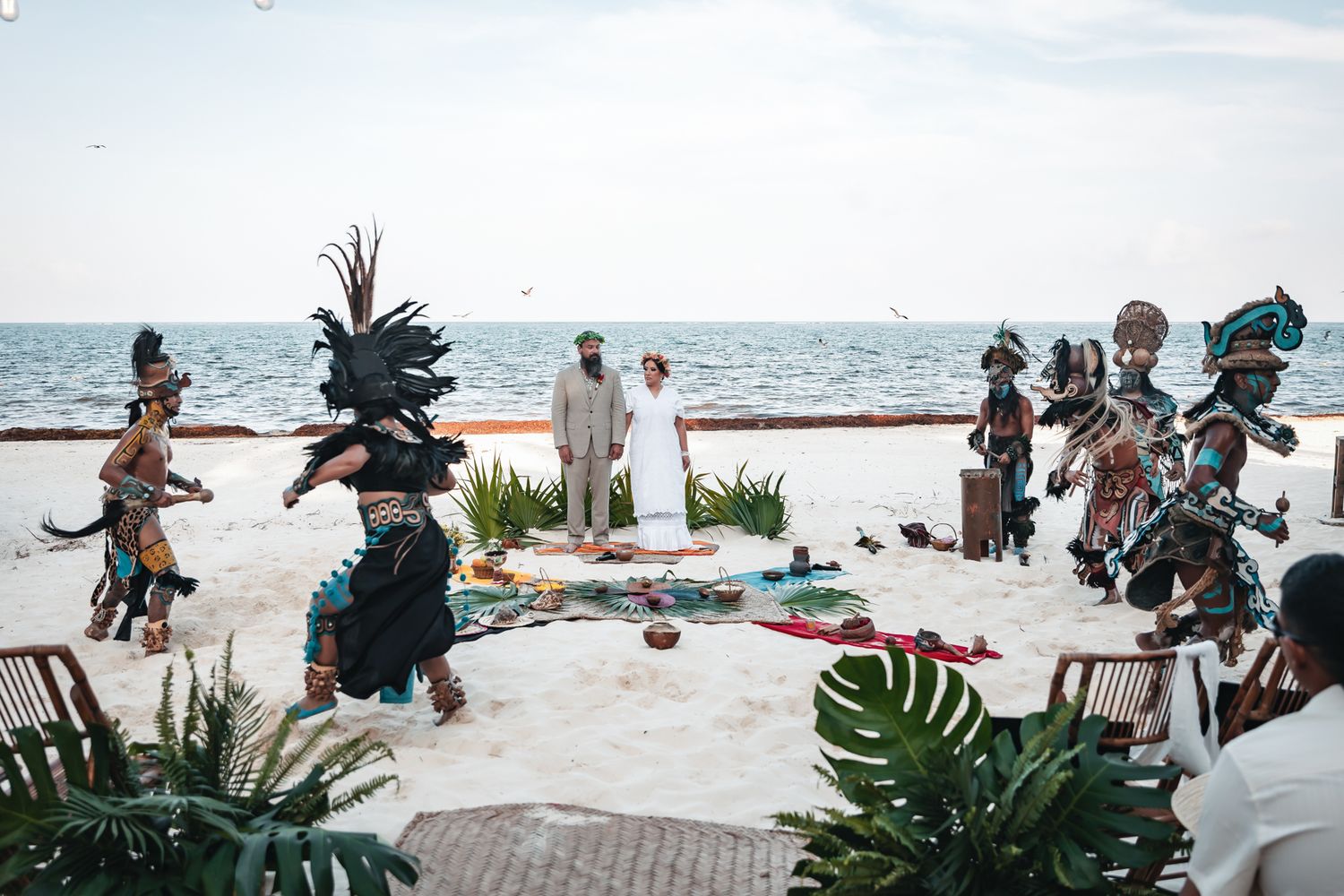Mayan dancers in traditional feathered costumes perform at a beachside wedding ceremony with ocean views.