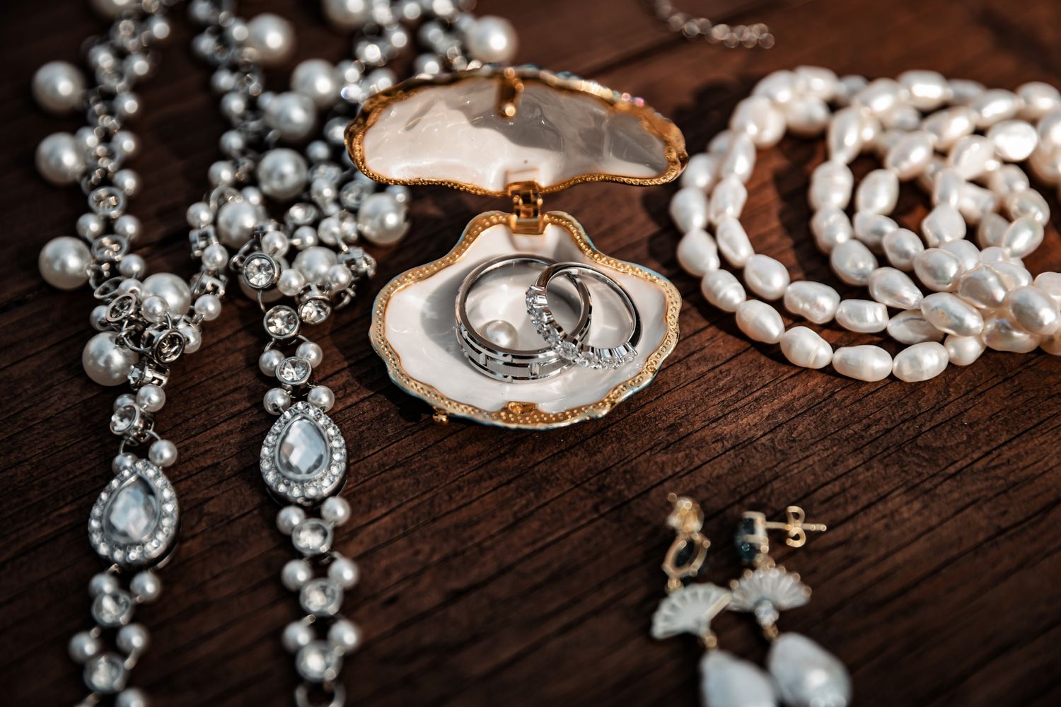 Elegant bridal jewelry flat lay featuring pearl necklaces, diamond drop earrings, and wedding rings in an oyster shell on wood.