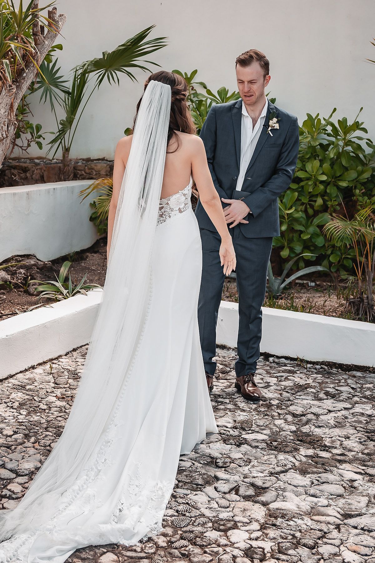 Bride in backless gown with veil and groom in dark suit share a first look on a cobblestone path.