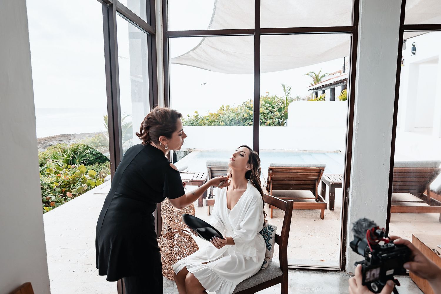 Makeup artist applying makeup to a woman in a white robe seated outdoors near a pool at a luxury resort.