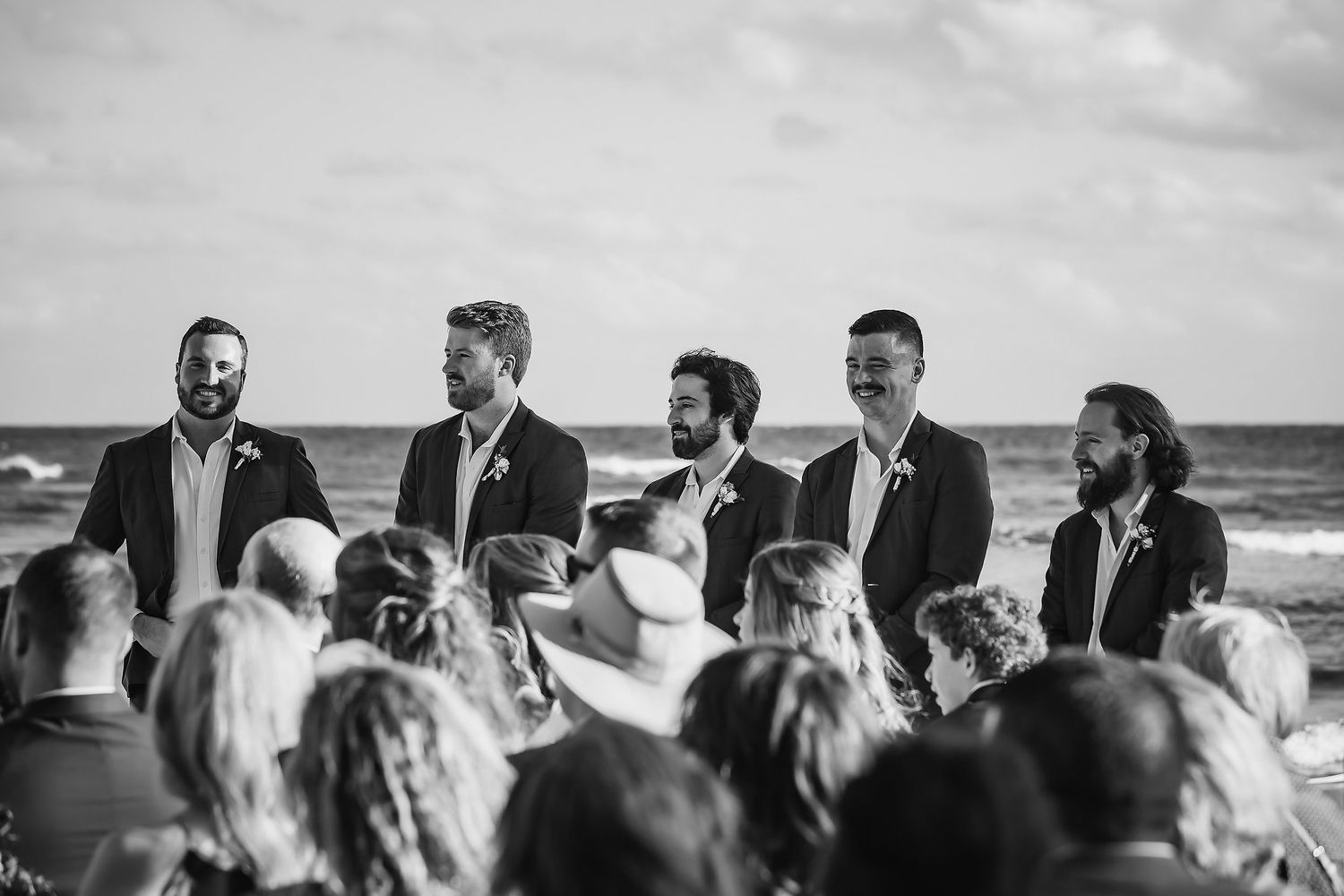 Black and white photo of groomsmen standing at an outdoor beach wedding ceremony with ocean waves in the background.