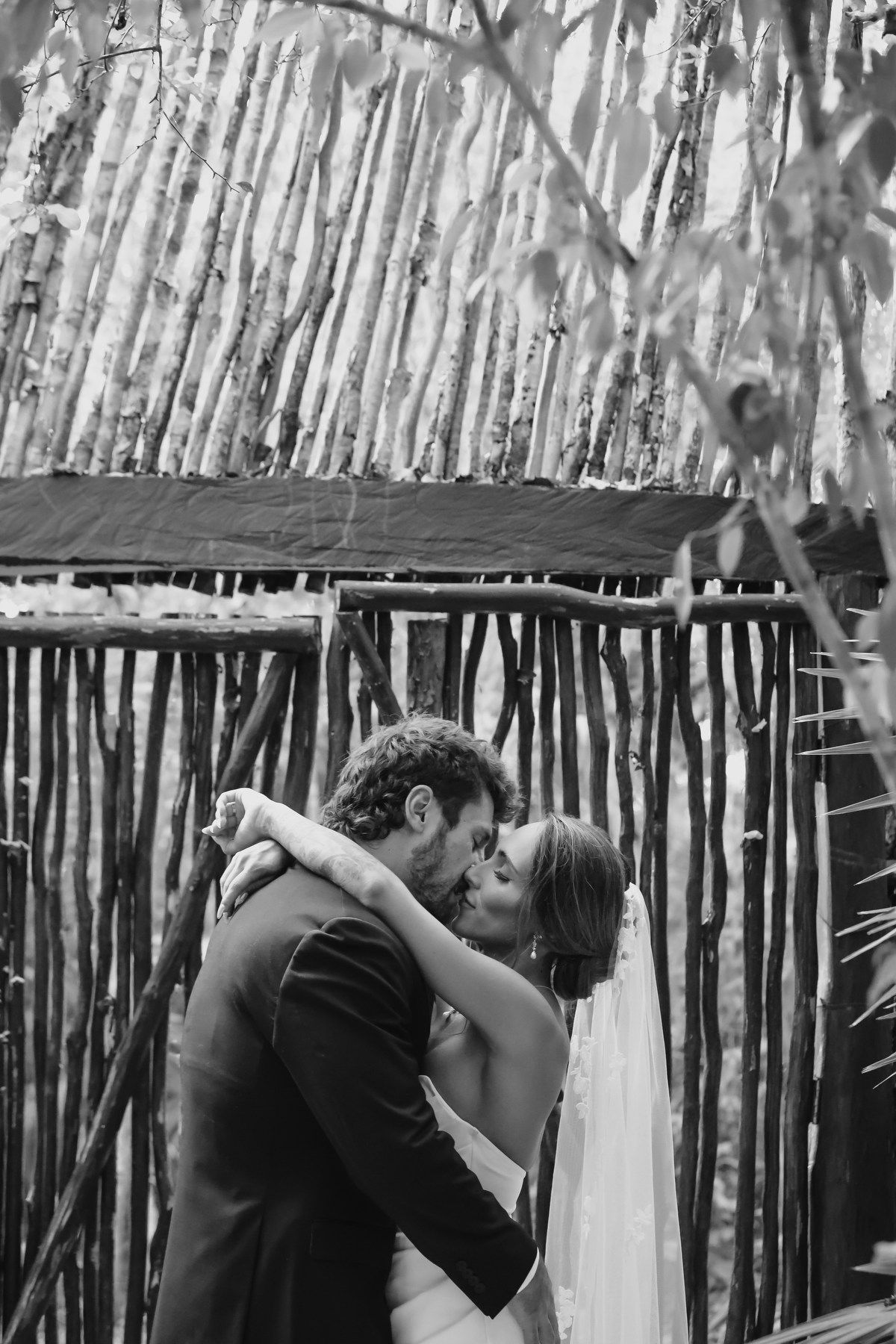 Black and white photo of a bride and groom sharing a kiss in front of a rustic wooden fence backdrop.