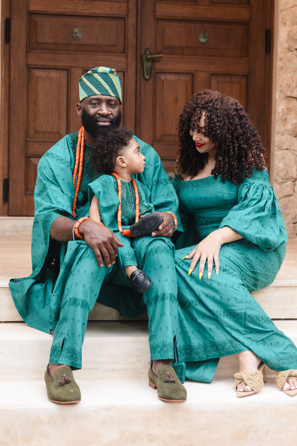 Family of three in matching teal traditional African attire sitting on steps, smiling warmly together.