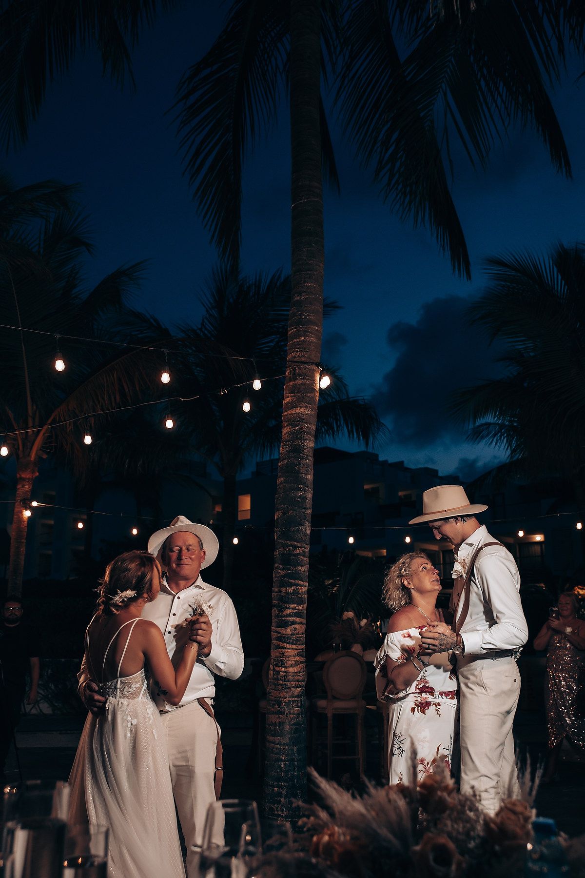 Couples dancing at a romantic beach wedding reception with string lights and palm trees at dusk.
