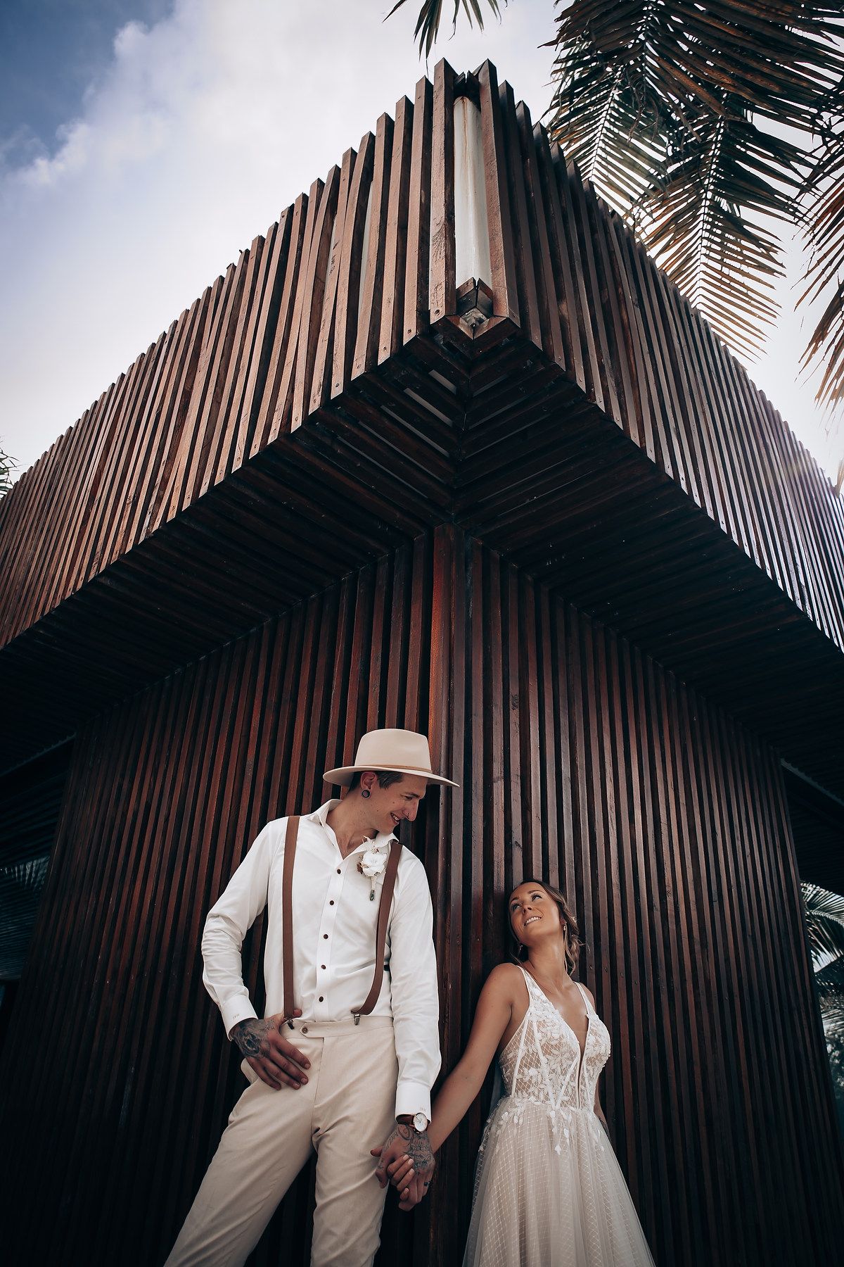 A stylish couple poses against modern wooden architecture with palm trees.