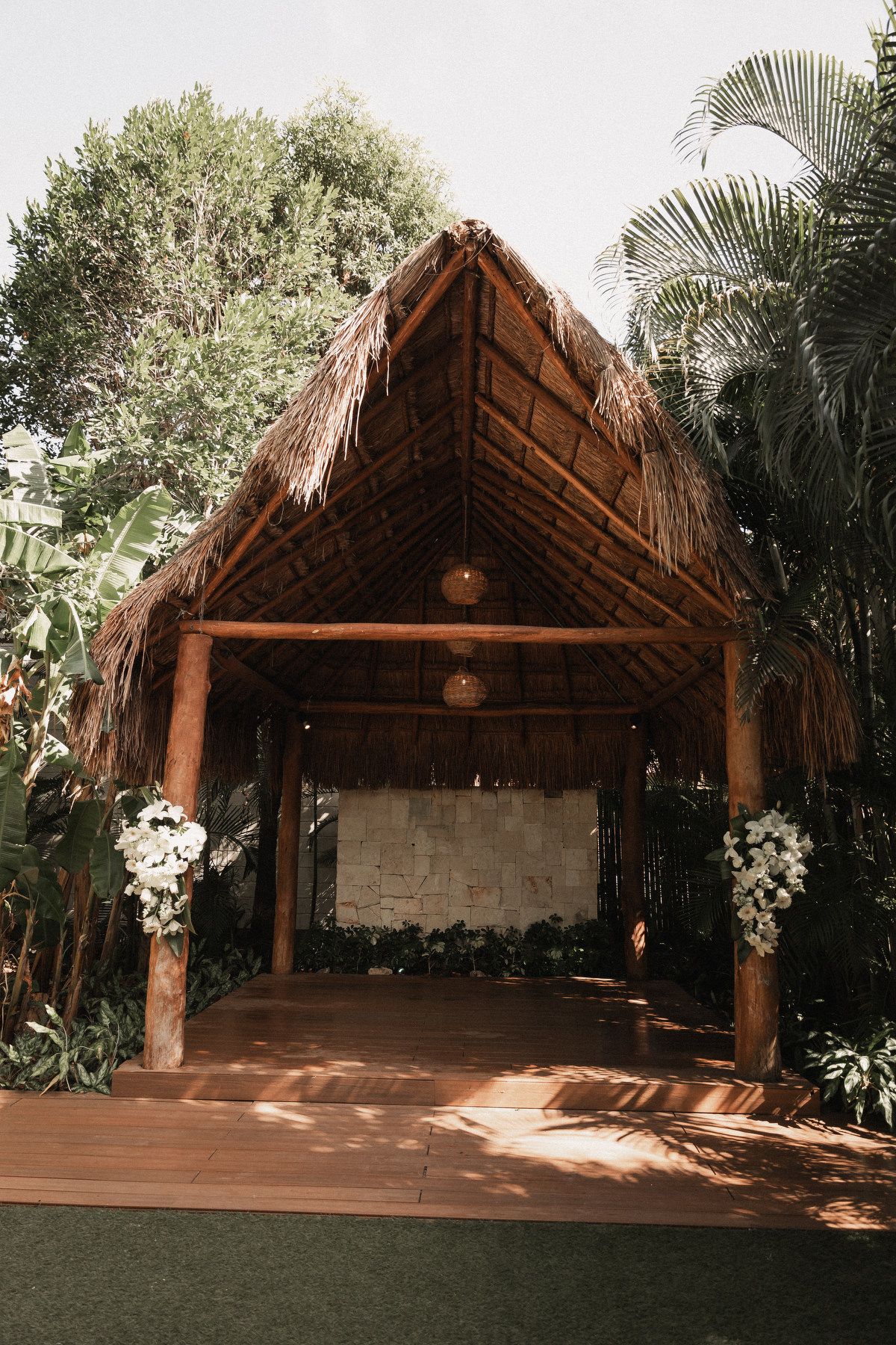 Rustic thatched-roof wedding ceremony altar decorated with white floral arrangements amid lush tropical greenery.