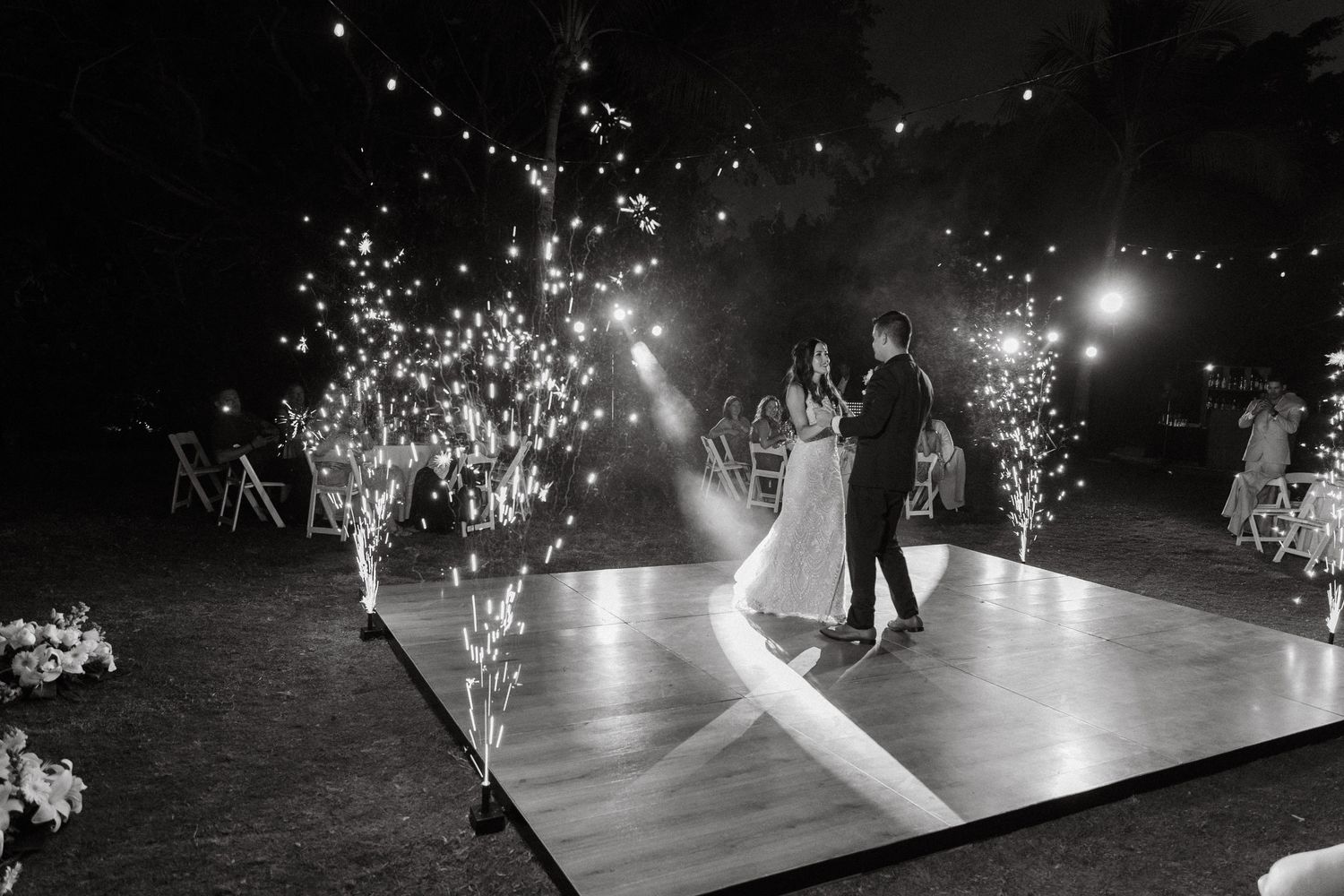 Newlyweds share first dance on illuminated floor with sparkling fireworks in black and white.
