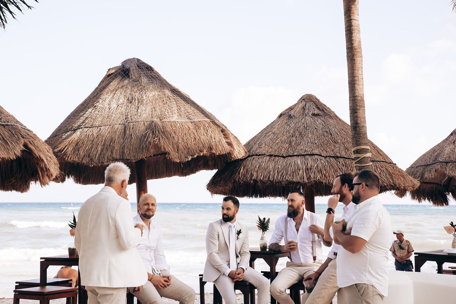 Wedding guests dancing and celebrating under thatched roof palapas at a beach resort during daytime.