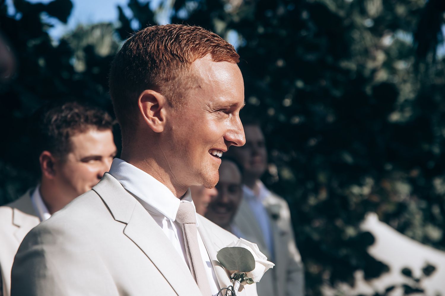 Groomsmen in light colored suits gather outdoors during a summer wedding celebration.