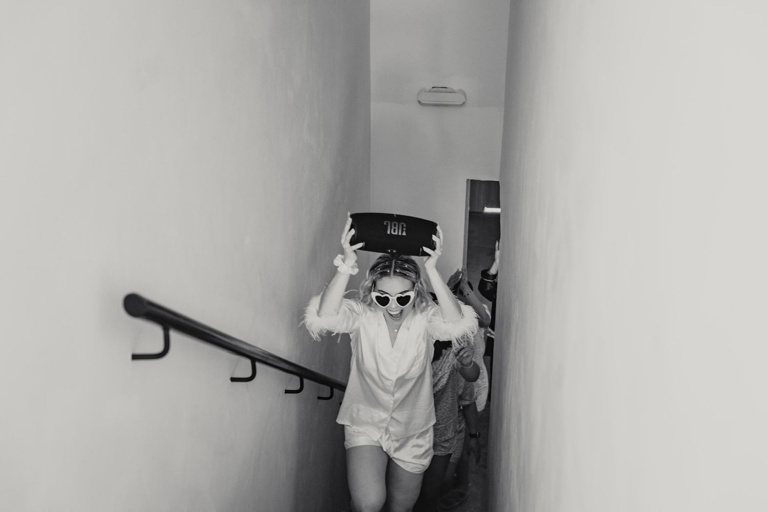 Black and white photo of a woman carrying a box on her head while walking up stairs in a white outfit and sunglasses.