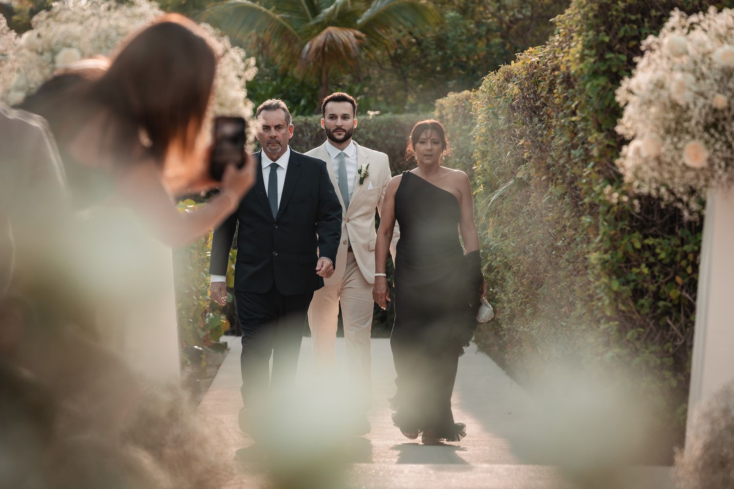 Groom in beige suit walking with guests through lush garden ceremony path.