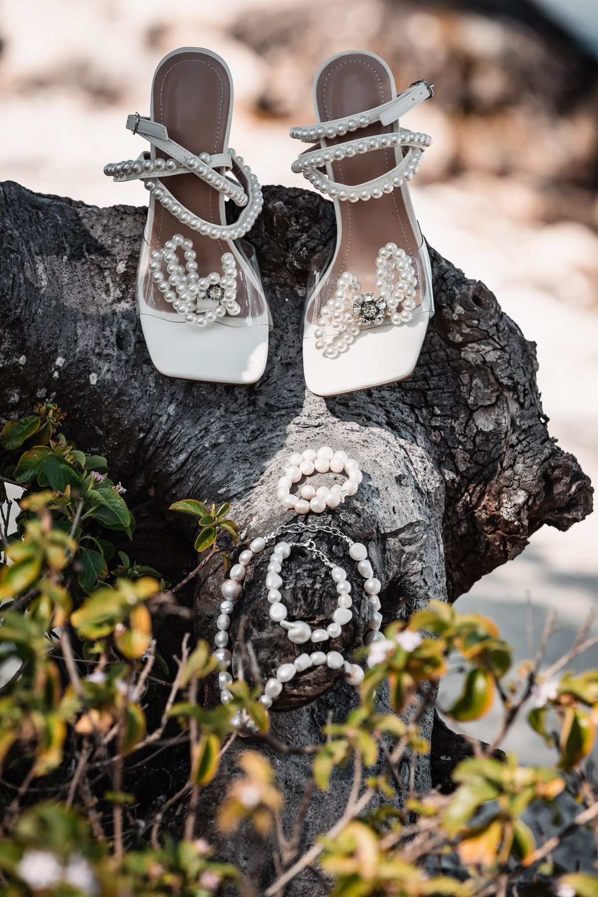 Elegant pearl-embellished white bridal heels and pearl bracelet displayed on driftwood outdoors.