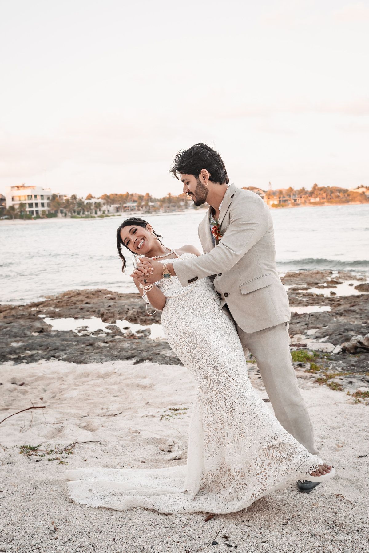 Bride and groom share a romantic dip dance on a rocky beach at sunset, bride in lace gown, groom in tan suit.