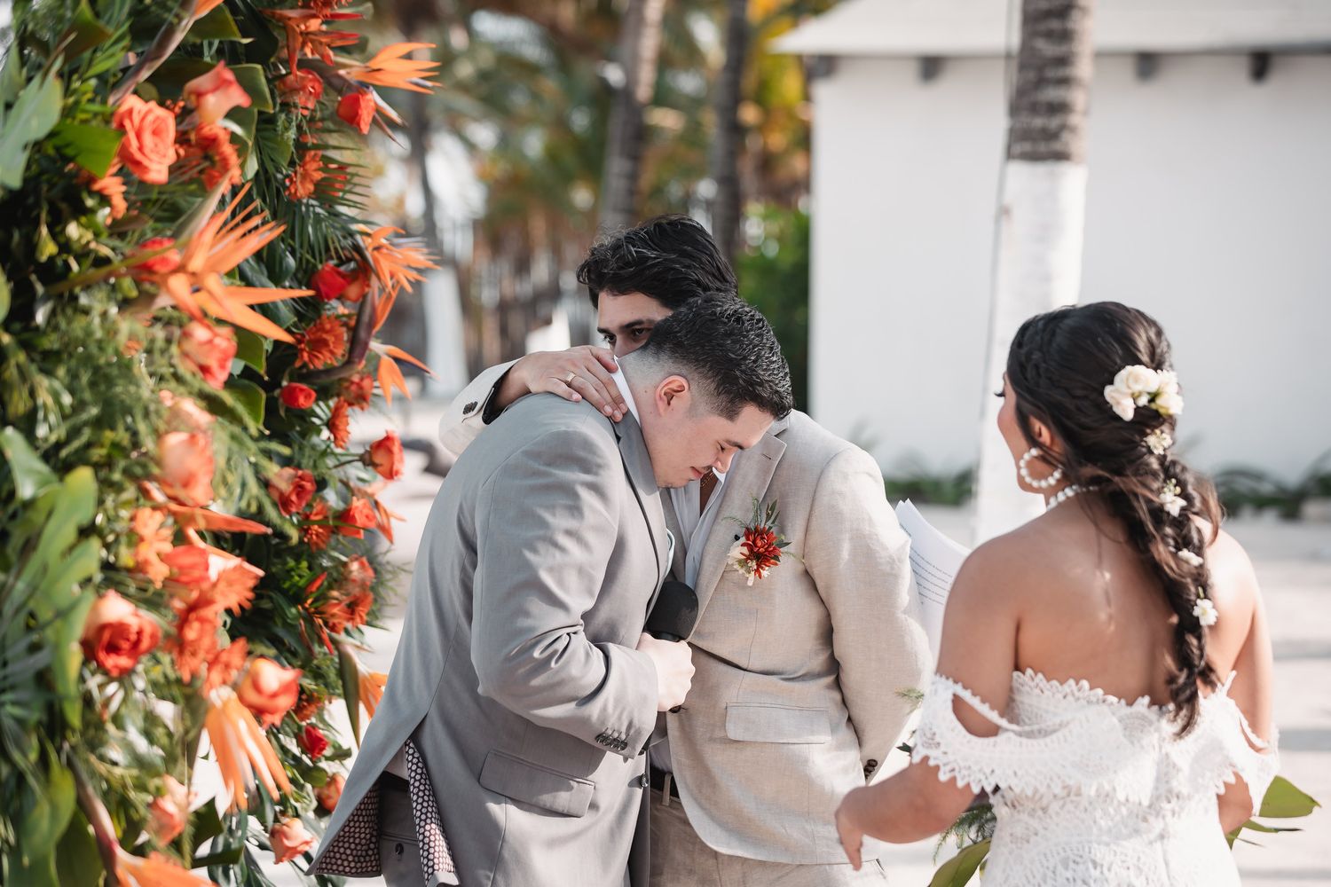Groom in gray suit shares emotional moment with guest at tropical floral wedding altar.