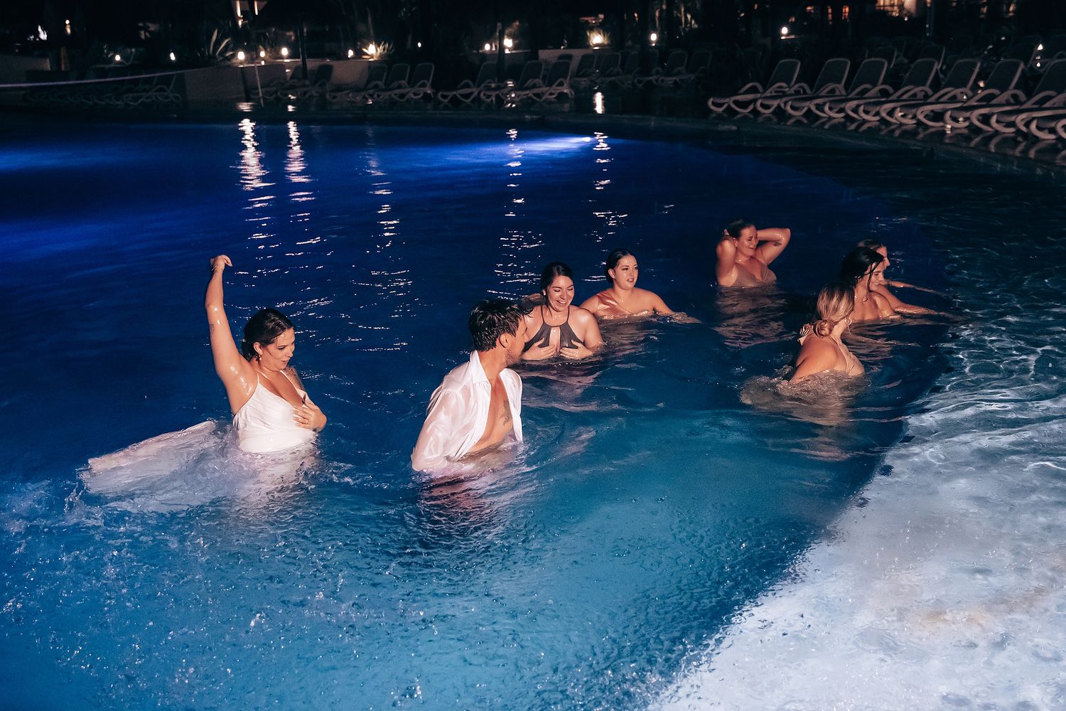 Wedding guests celebrate in a blue-lit swimming pool at night.