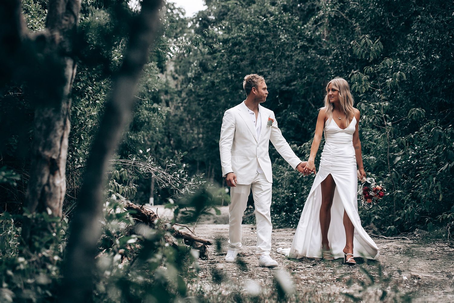 A couple in elegant white outfits pose together on a forest path during their wedding.