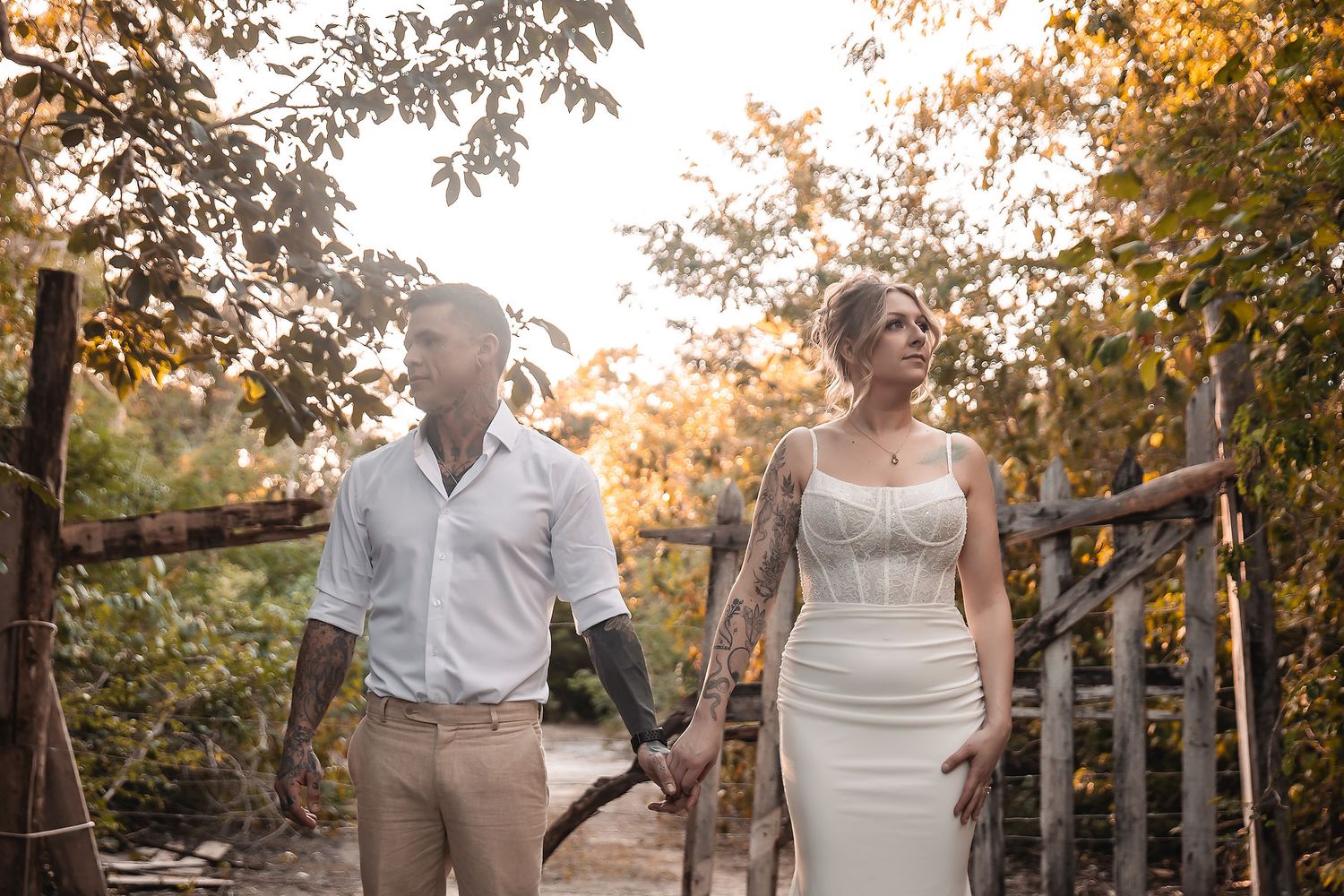 Couple in wedding attire holding hands outdoors among autumn trees, bride in white gown, groom in casual white shirt.