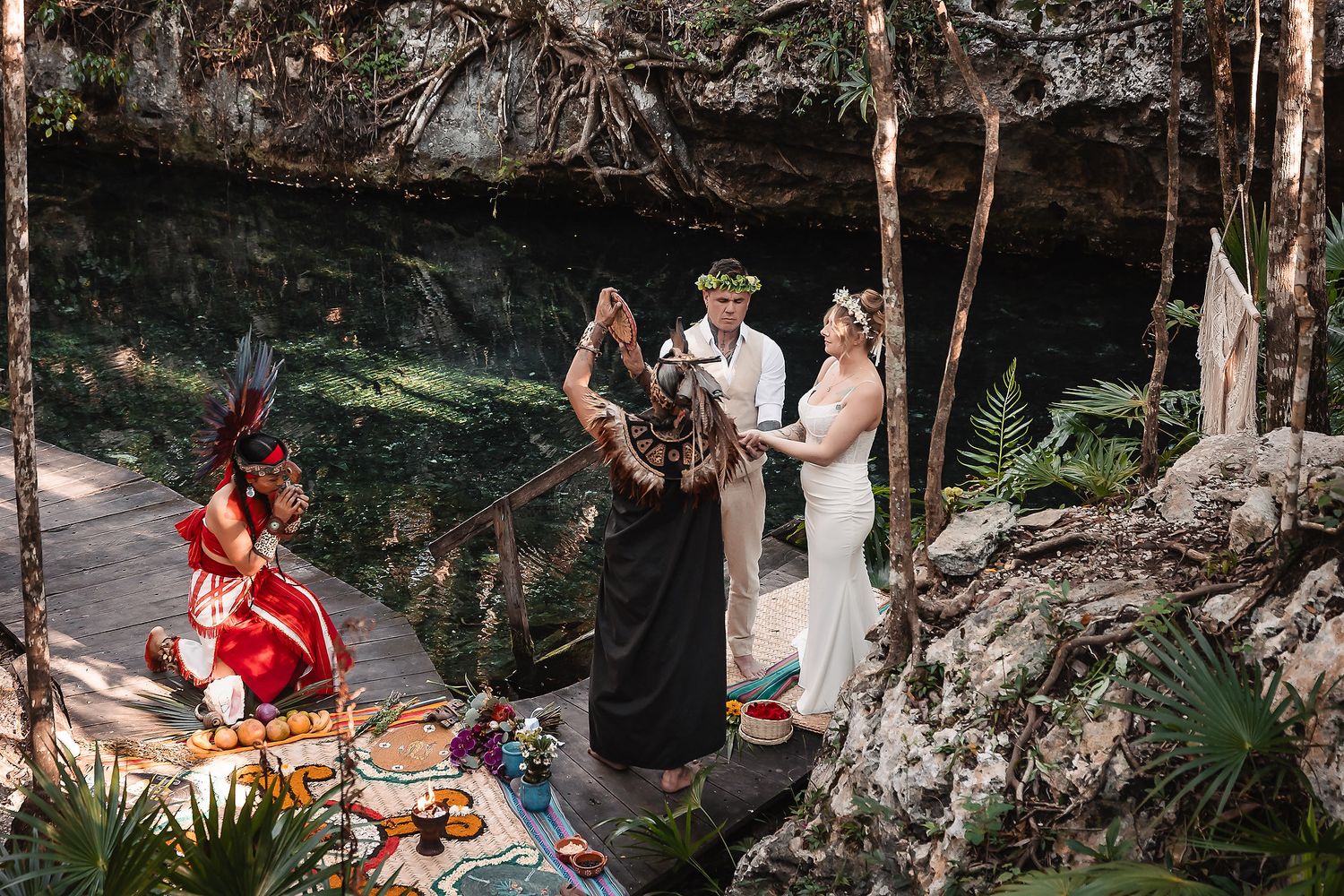 Mayan wedding ceremony in a lush cenote jungle setting with officiant, bride in white dress, and guest in red attire.