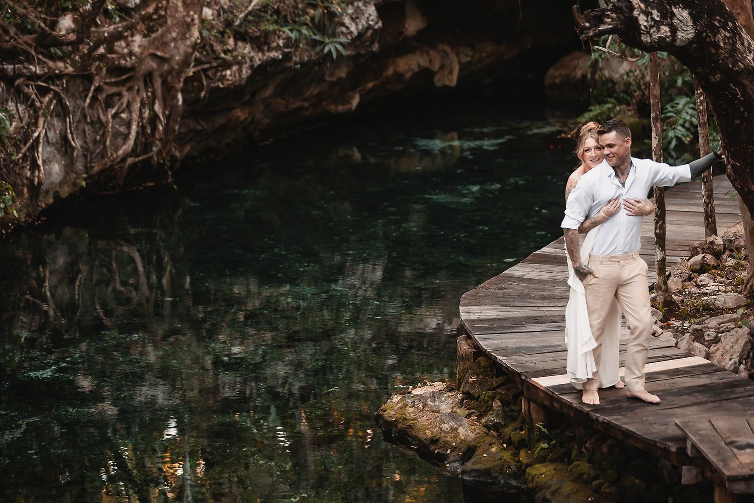 Couple embracing on wooden boardwalk over turquoise cenote waters surrounded by lush tropical cave vegetation.