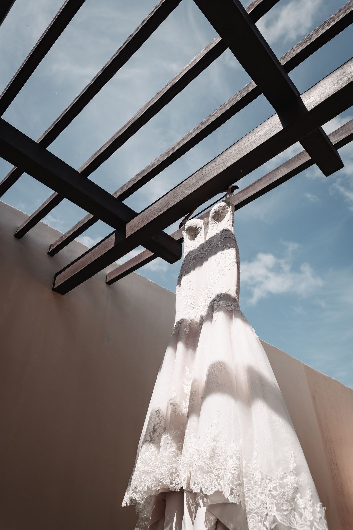 White lace wedding dress hanging under a wooden pergola against a blue sky.