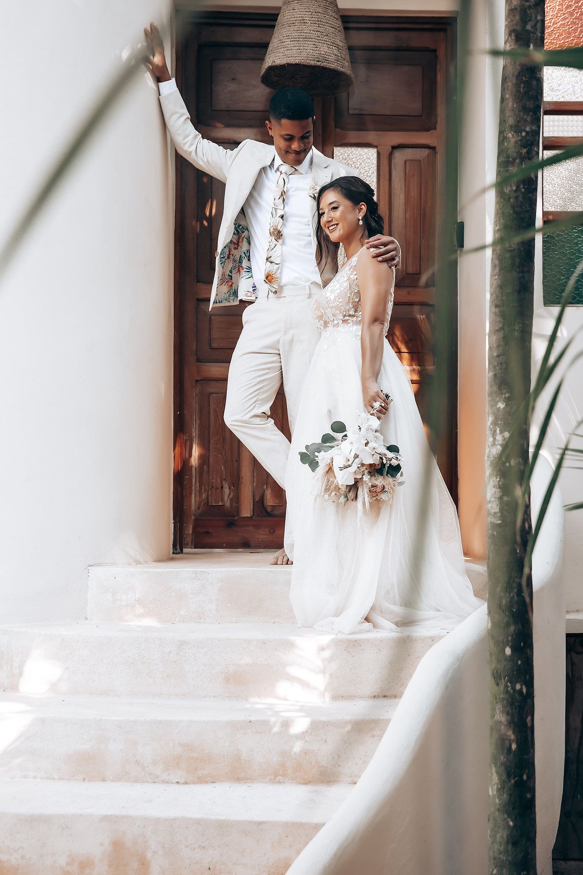 Wedding couple in elegant white attire posing on outdoor steps of a bright building.
