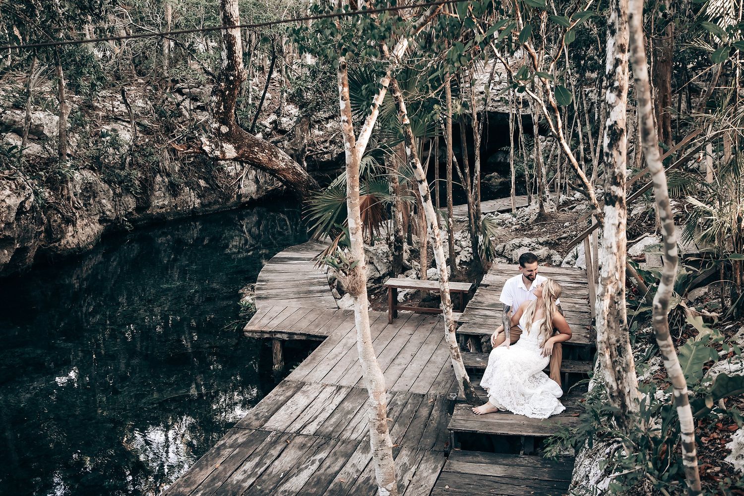 A wooden boardwalk leads to a tranquil cenote surrounded by lush tropical vegetation.