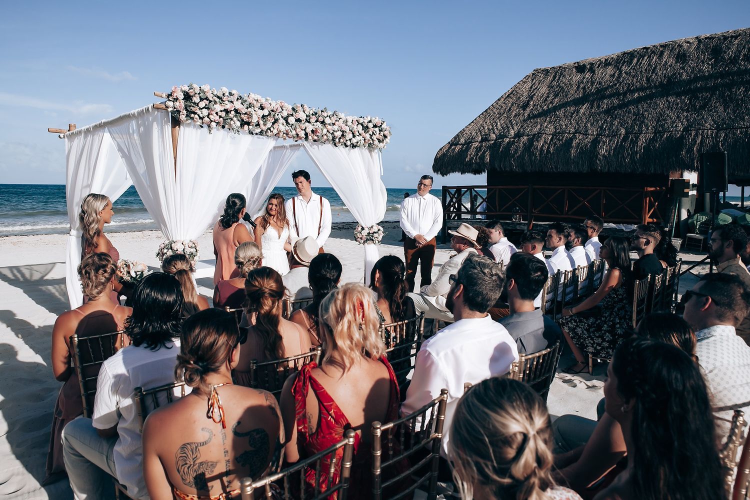 Beach wedding ceremony setup with white canopy and gathered guests on sandy shores.