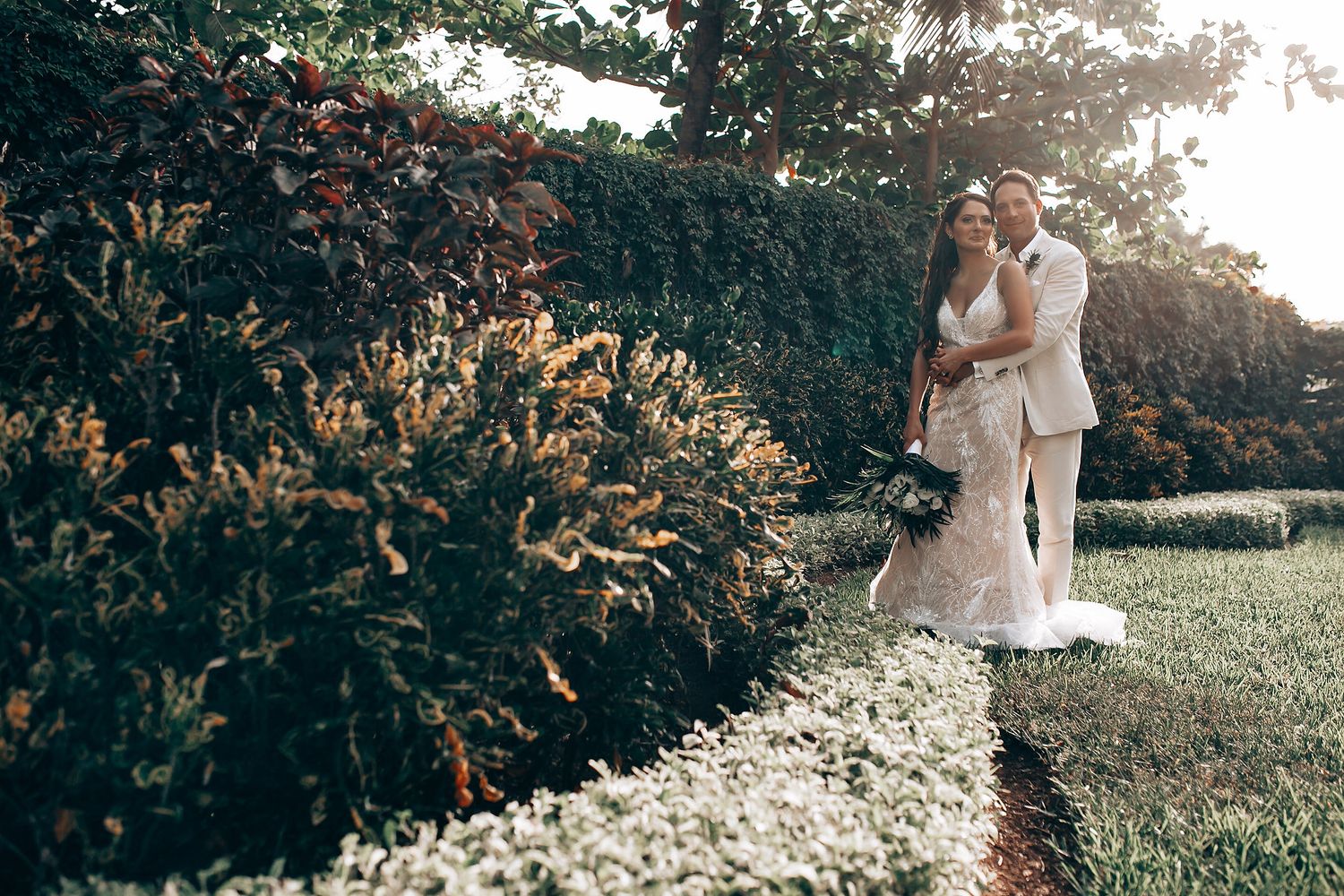 Romantic wedding couple embracing in a sunlit garden with white flowers.