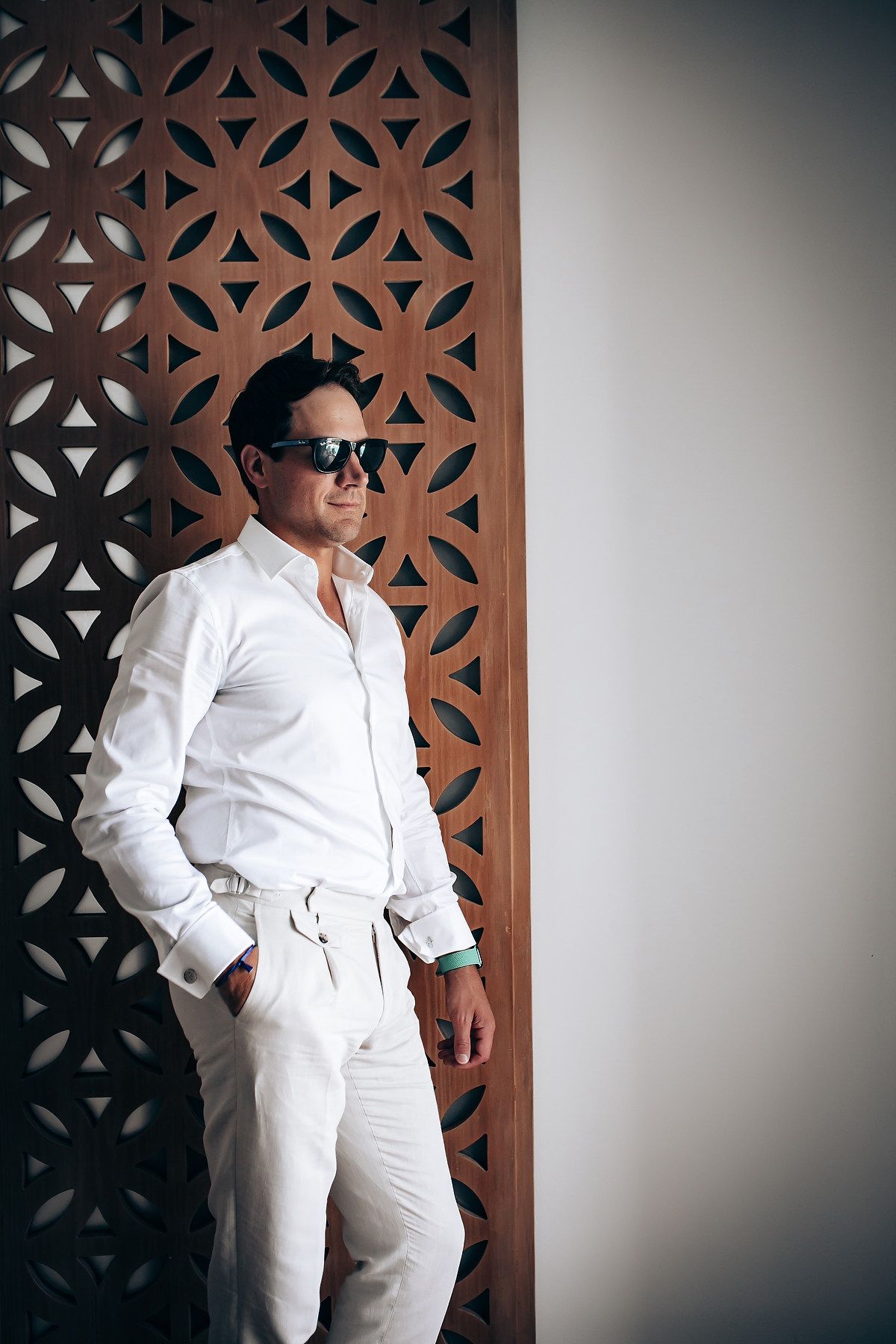 A person in an all-white outfit and sunglasses poses against a decorative geometric wall pattern.