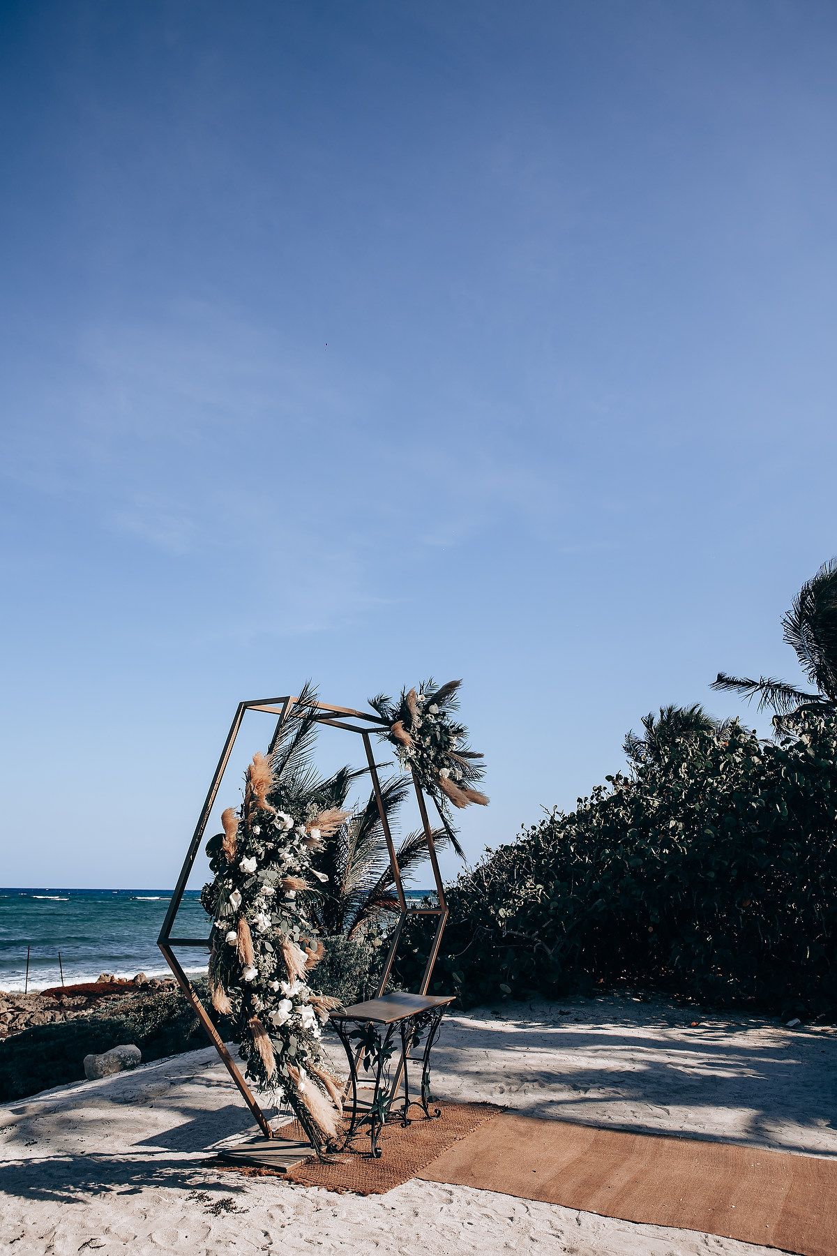 Beach wedding ceremony setup with wooden arch and floral decorations against ocean backdrop.