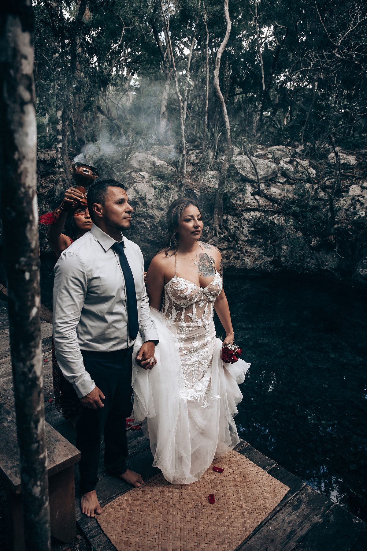 A couple in wedding attire sits together on a wooden platform in a moody forest setting.