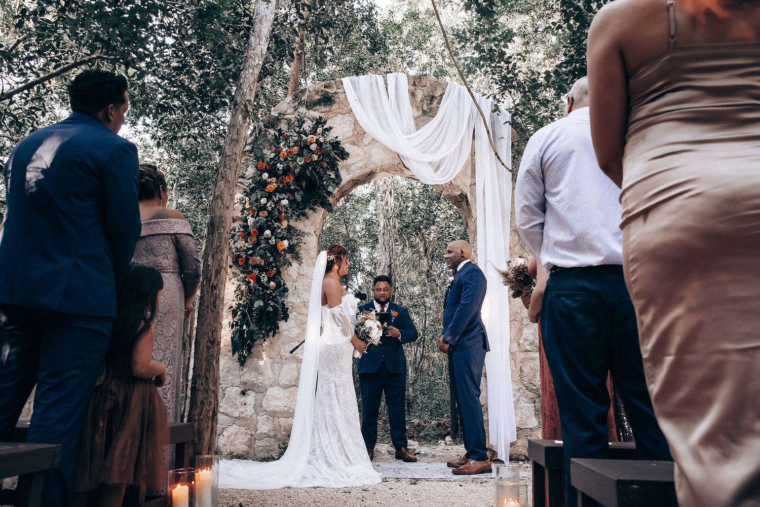 Outdoor wedding ceremony with guests gathered around stone archway.