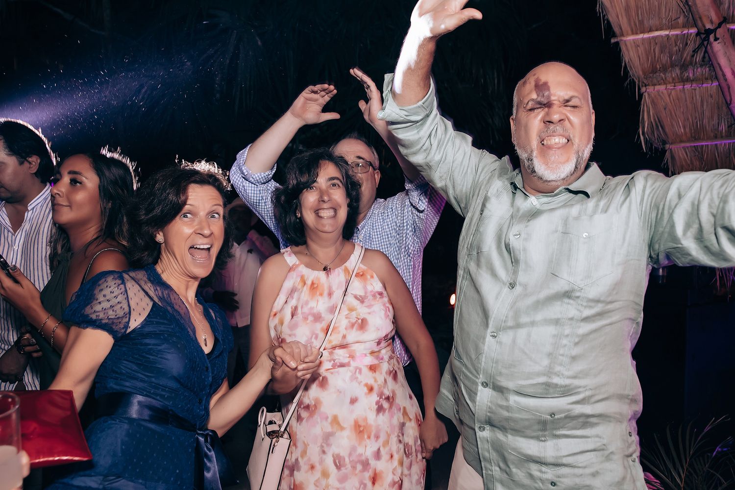 Joyful wedding guests dance and celebrate with raised arms on a dimly lit dance floor.