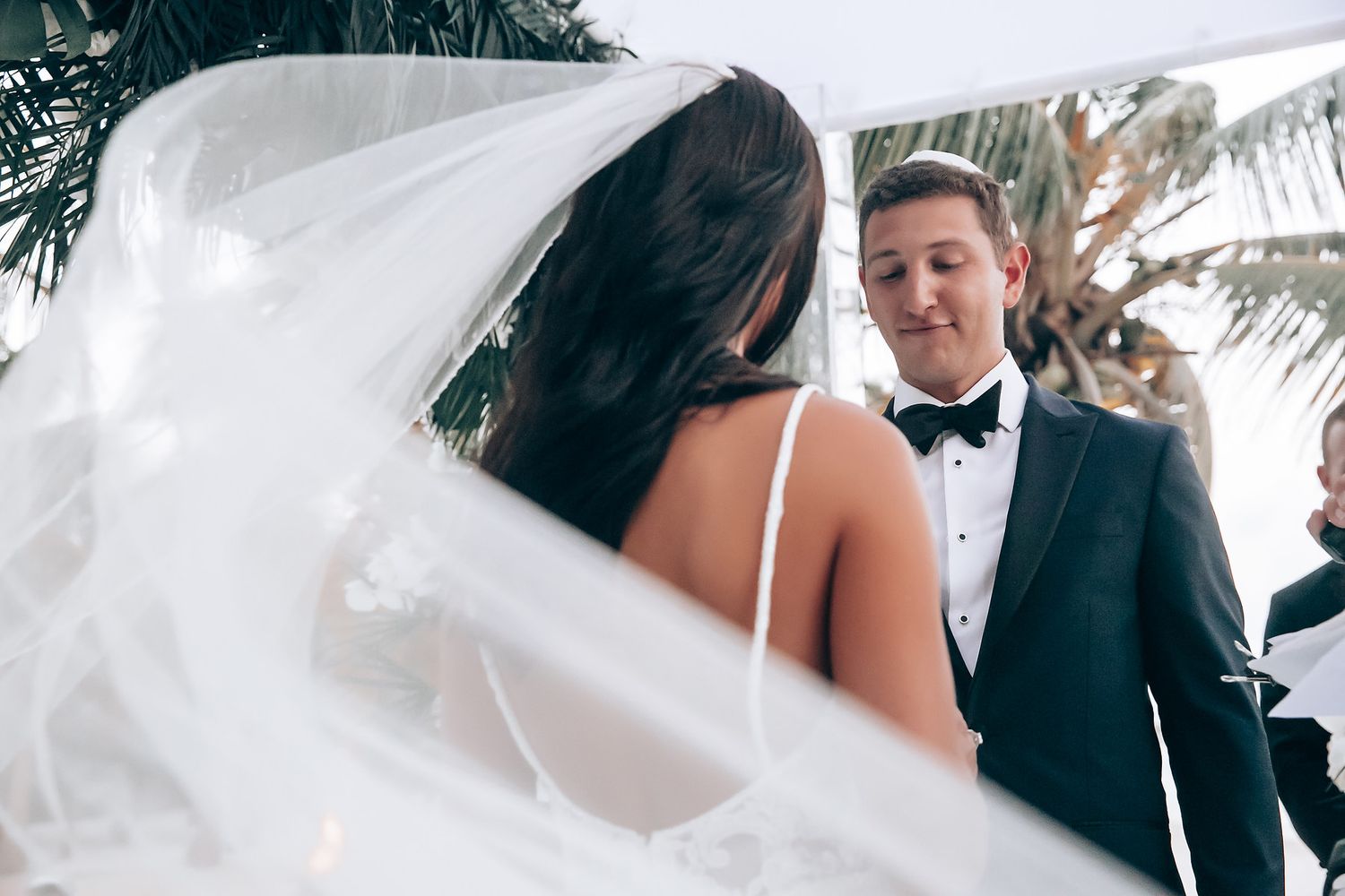 Romantic beach wedding moment with flowing white veil under tropical palms.