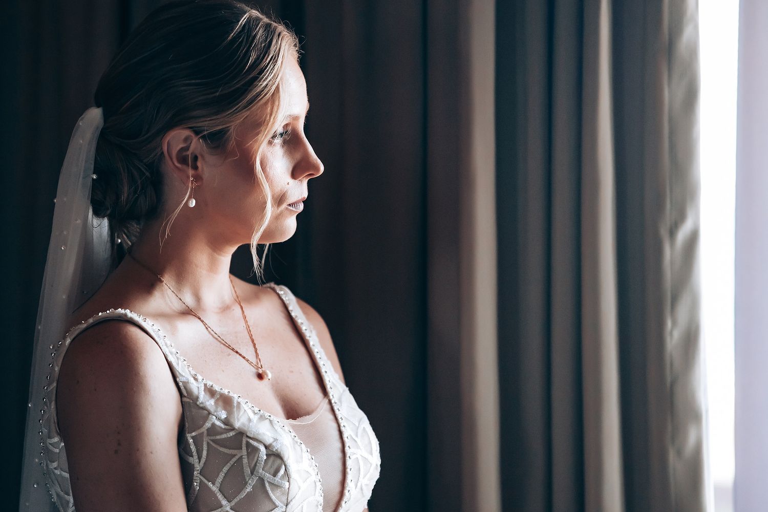 A woman in a wedding dress stands by a window, looking pensive.