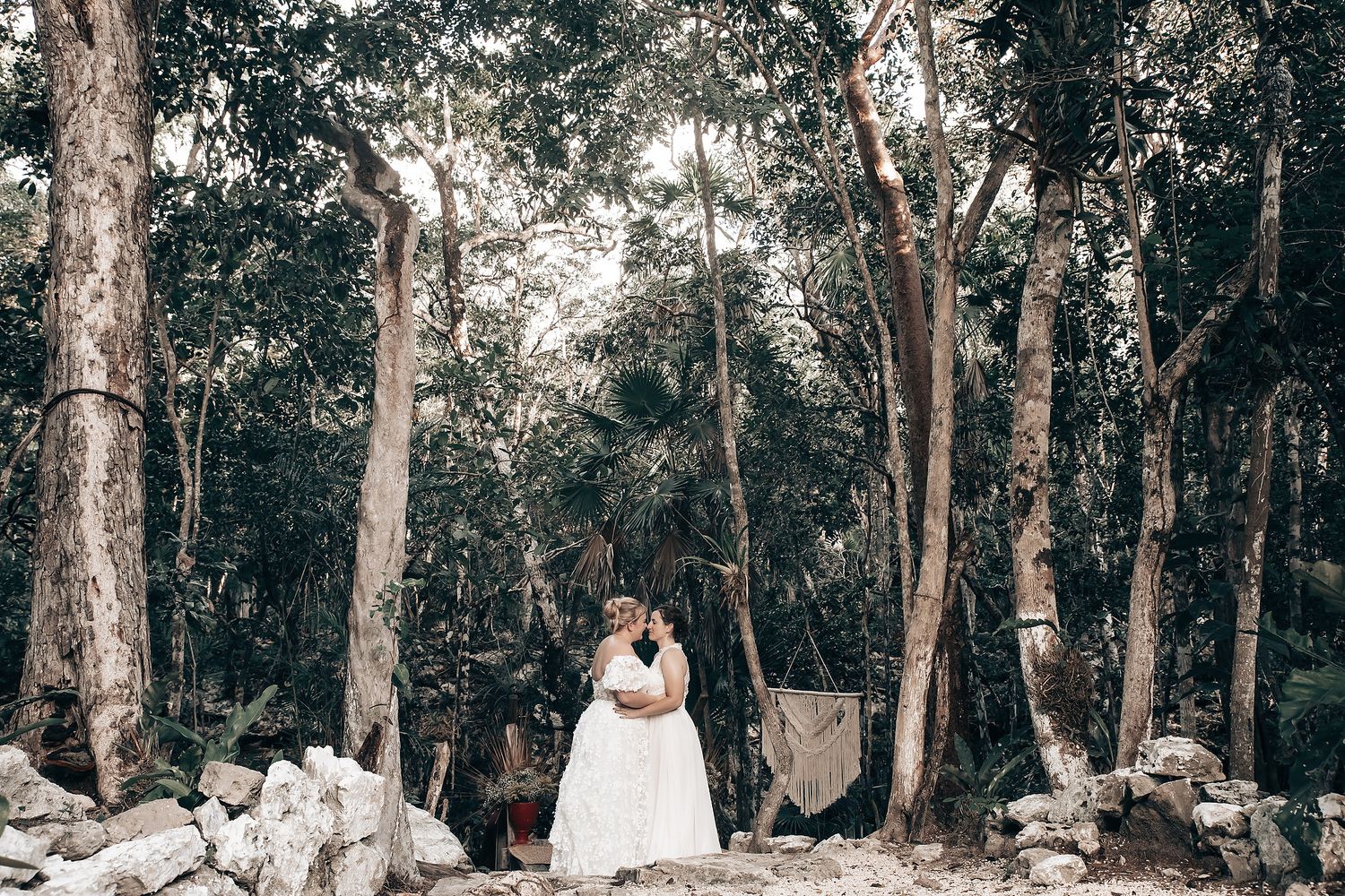 Two brides in white wedding dresses standing together with a person in traditional indigenous attire.